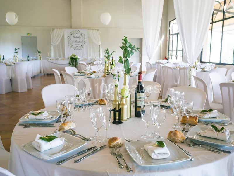 Salle de réception décorée pour un événement, avec tables rondes dressées, nappes blanches et éléments végétaux.
