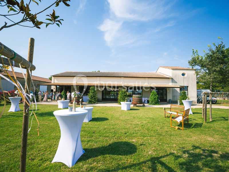 Extérieur d'une salle de réception avec pelouse, tables hautes drapées de blanc et quelques invités en arrière-plan.