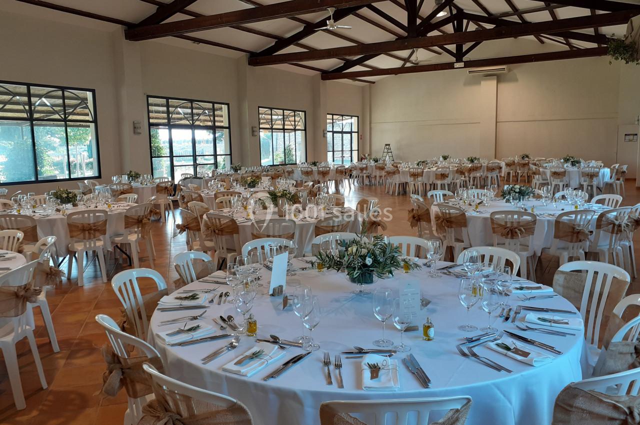 Salle de réception lumineuse avec tables rondes décorées pour un événement, chaises blanches et poutres apparentes au…