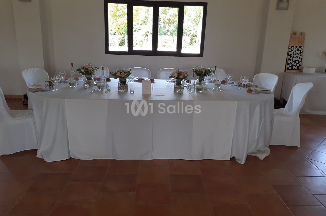 Table rectangulaire décorée pour un repas, avec nappes blanches, fleurs et vaisselle, dans une salle lumineuse.