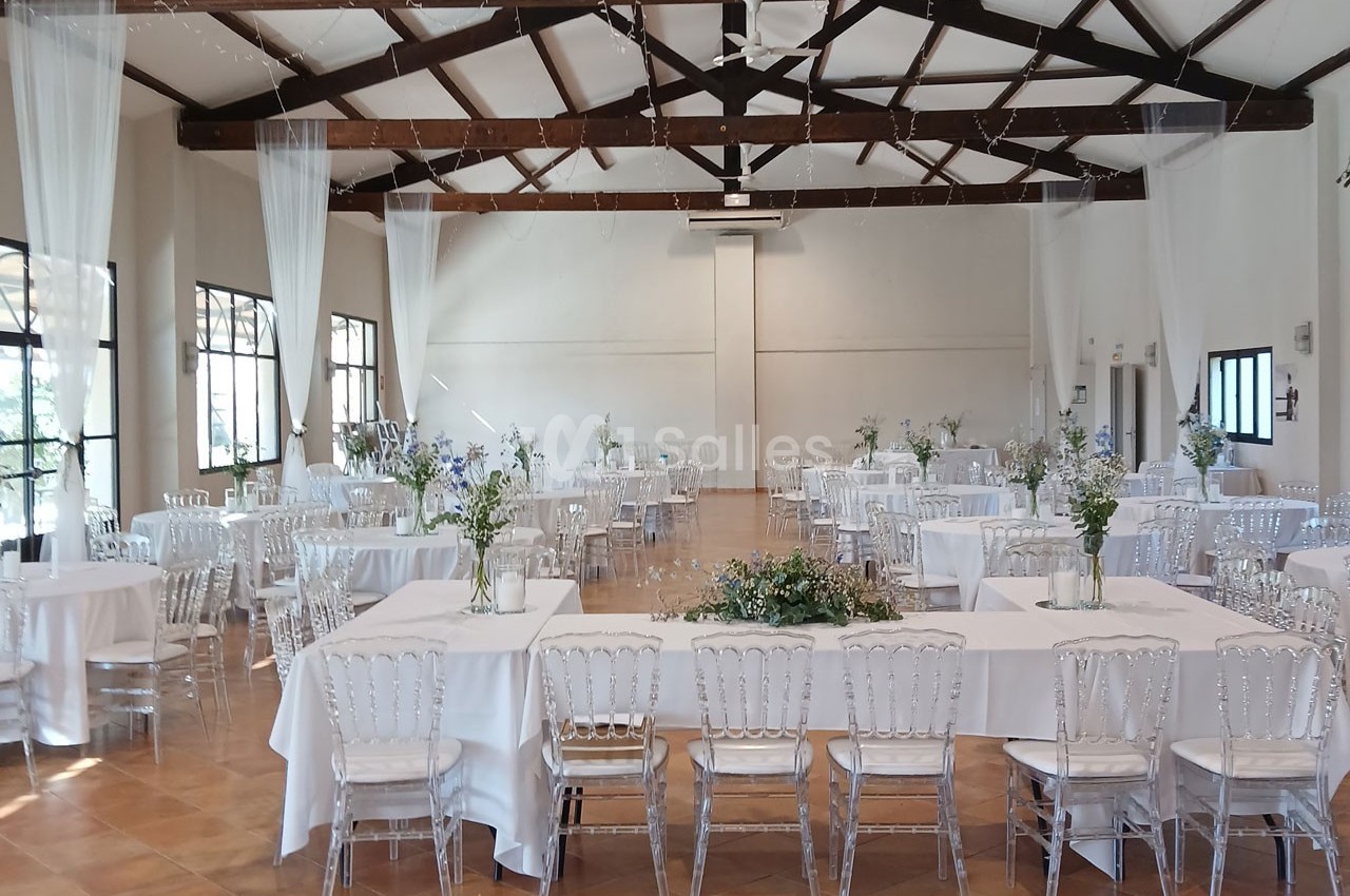 Salle de réception lumineuse avec tables dressées, nappes blanches, chaises transparentes et décorations florales.