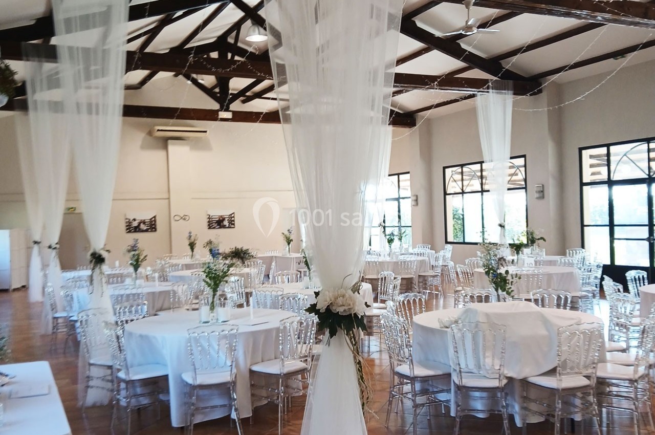 Salle de réception lumineuse décorée pour un événement, avec tables rondes, chaises transparentes et voilages suspendus.