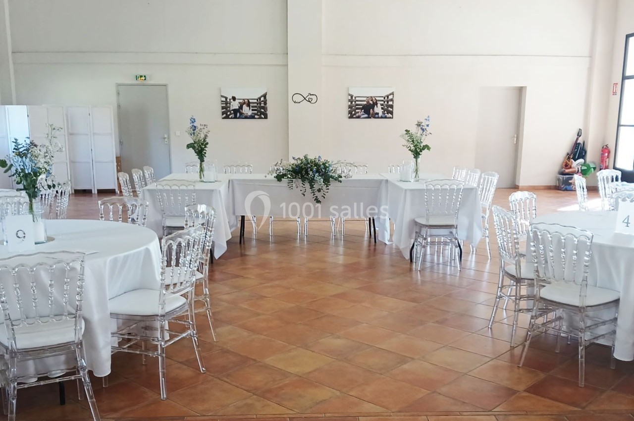Salle de réception lumineuse avec tables rondes dressées de nappes blanches, chaises transparentes et décorations florales.