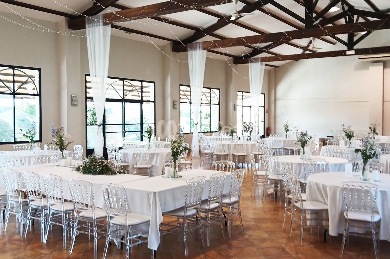 Salle de réception lumineuse avec tables rondes dressées, chaises transparentes et décorations florales.