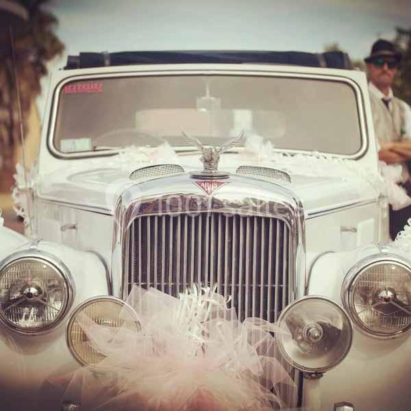 Voiture ancienne blanche décorée de rubans roses, probablement pour un mariage, avec un homme en arrière-plan.