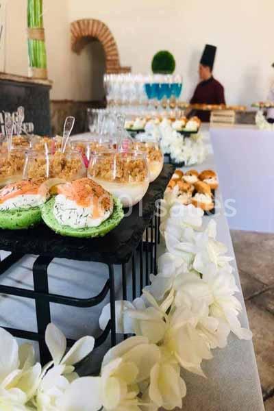 Plateau de verrines et amuse-bouches disposés sur une table décorée de fleurs blanches, avec un chef en arrière-plan.