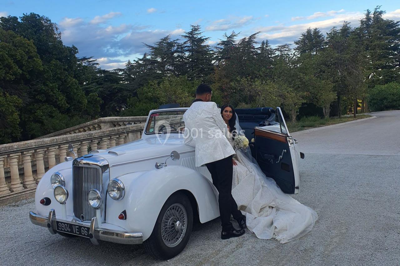 Un couple en tenue de mariage pose près d'une voiture ancienne blanche, dans un cadre extérieur arboré.