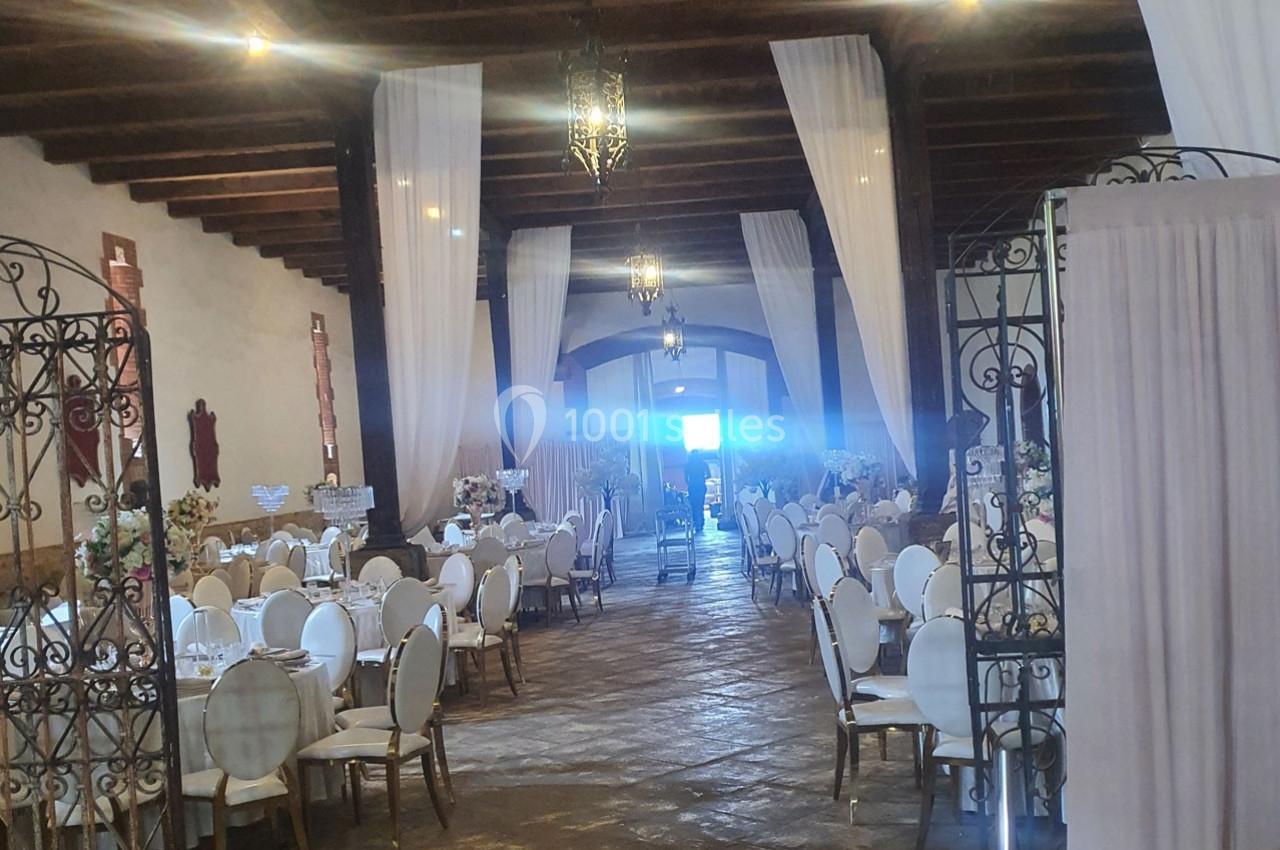 Salle de réception élégante avec tables dressées, chaises blanches, rideaux translucides et lumière naturelle au fond.