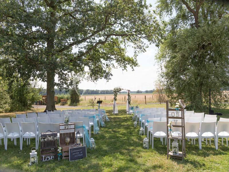 Location salle La Chapelle-Gauthier (Seine-et-Marne) - Nomade-Lodge #14 Chaises blanches alignées en extérieur sous des arbres, formant une allée menant à une arche de mariage décorée.