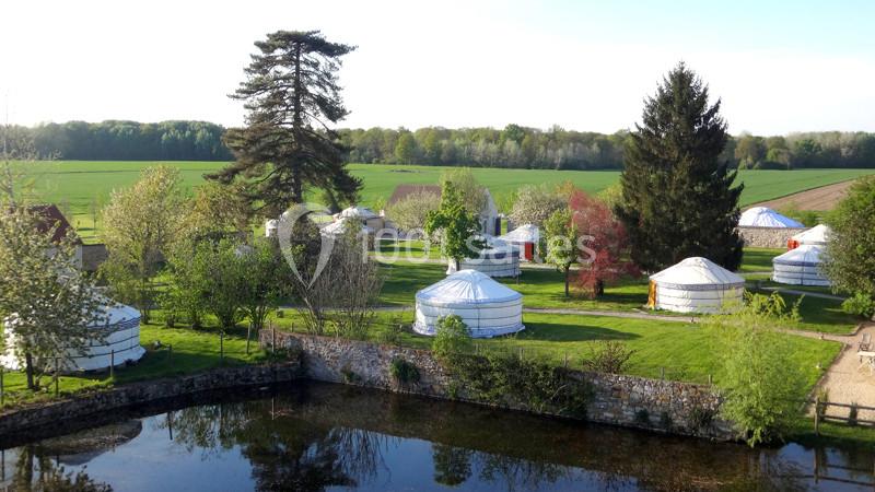 Location salle La Chapelle-Gauthier (Seine-et-Marne) - Nomade-Lodge #13 Vue d'un ensemble de yourtes blanches installées dans un paysage verdoyant avec des arbres et un étang au premier plan.