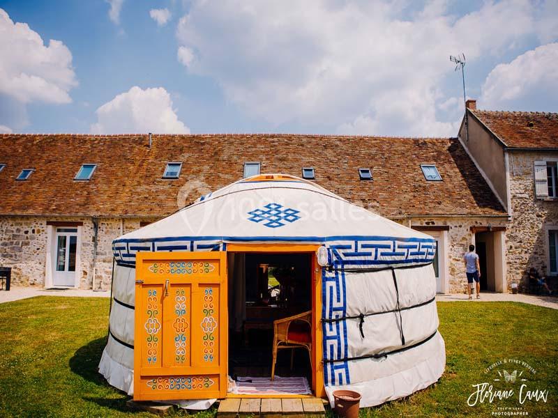Location salle La Chapelle-Gauthier (Seine-et-Marne) - Nomade-Lodge #15 Yourte blanche décorée de motifs bleus et orange, installée devant une maison en pierre sous un ciel partiellement nuageux.