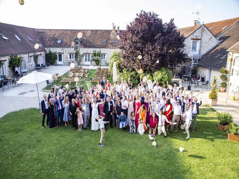 Location salle La Chapelle-Gauthier (Seine-et-Marne) - Nomade-Lodge #16 Groupe de personnes rassemblées dans une cour verdoyante devant des bâtiments en pierre, levant les bras en l'air.