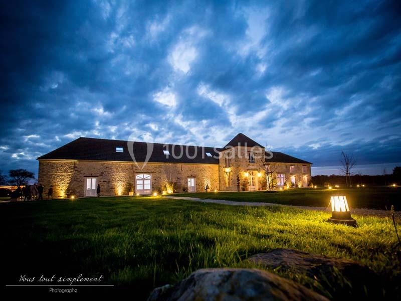 Location salle La Chapelle-Gauthier (Seine-et-Marne) - Nomade-Lodge #17 Bâtiment en pierre éclairé à la tombée de la nuit, entouré d'une pelouse et d'un ciel nuageux.