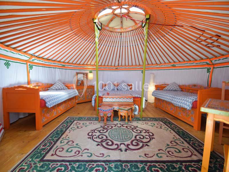 Location salle La Chapelle-Gauthier (Seine-et-Marne) - Nomade-Lodge #18 Intérieur d'une yourte colorée avec des lits en bois, un tapis orné et une table basse au centre.