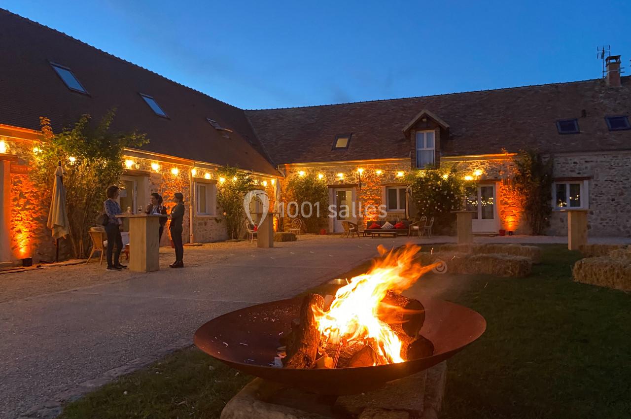 Location salle La Chapelle-Gauthier (Seine-et-Marne) - Nomade-Lodge #19 Brasero allumé au premier plan dans une cour de ferme éclairée en soirée, avec deux personnes discutant près de tables…
