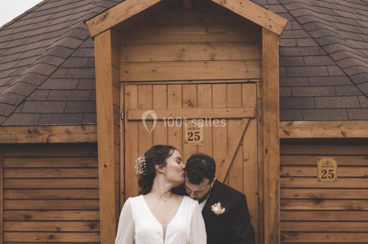 Location salle La Chapelle-Gauthier (Seine-et-Marne) - Nomade-Lodge #28 Un couple en tenue de mariage pose devant une petite cabane en bois avec un toit en pente.