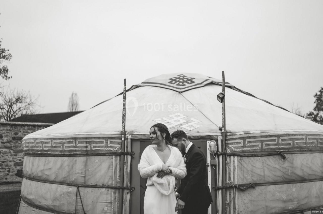 Location salle La Chapelle-Gauthier (Seine-et-Marne) - Nomade-Lodge #30 Un couple en tenue de mariage se tient devant une yourte dans un décor extérieur sobre et nuageux.