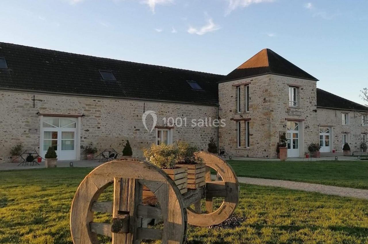 Location salle La Chapelle-Gauthier (Seine-et-Marne) - Nomade-Lodge #38 Bâtiment en pierre avec toit en ardoise, jardin verdoyant et grandes roues en bois décoratives au premier plan.