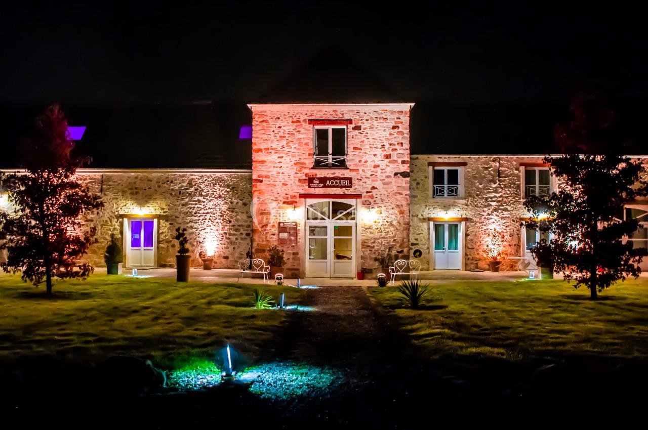 Location salle La Chapelle-Gauthier (Seine-et-Marne) - Nomade-Lodge #40 Façade d'un bâtiment en pierre éclairé par des lumières colorées, entouré d'un jardin la nuit.