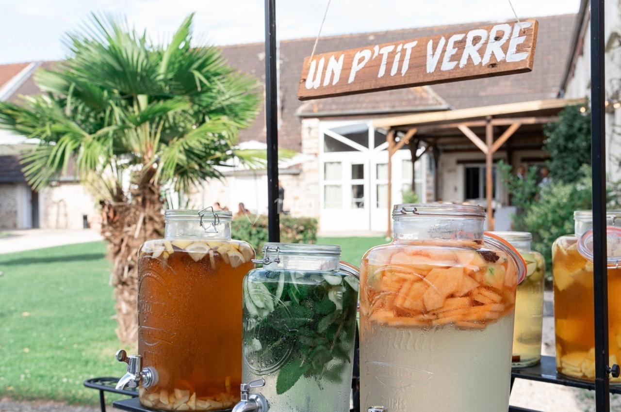 Location salle La Chapelle-Gauthier (Seine-et-Marne) - Nomade-Lodge #29 Des bonbonnes de boissons fruitées et infusées exposées en extérieur devant un bâtiment avec une enseigne ’Un p'tit verre’.