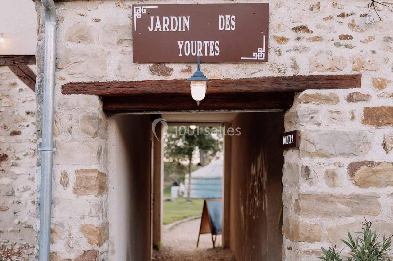 Location salle La Chapelle-Gauthier (Seine-et-Marne) - Nomade-Lodge #37 Entrée voûtée en pierre avec une enseigne ’Jardin des Yourtes’, menant à un espace extérieur avec une yourte visible.