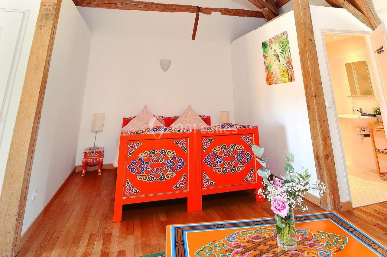 Location salle La Chapelle-Gauthier (Seine-et-Marne) - Nomade-Lodge #43 Lit en bois peint de motifs colorés dans une chambre lumineuse avec poutres apparentes et bouquet de fleurs sur une table.