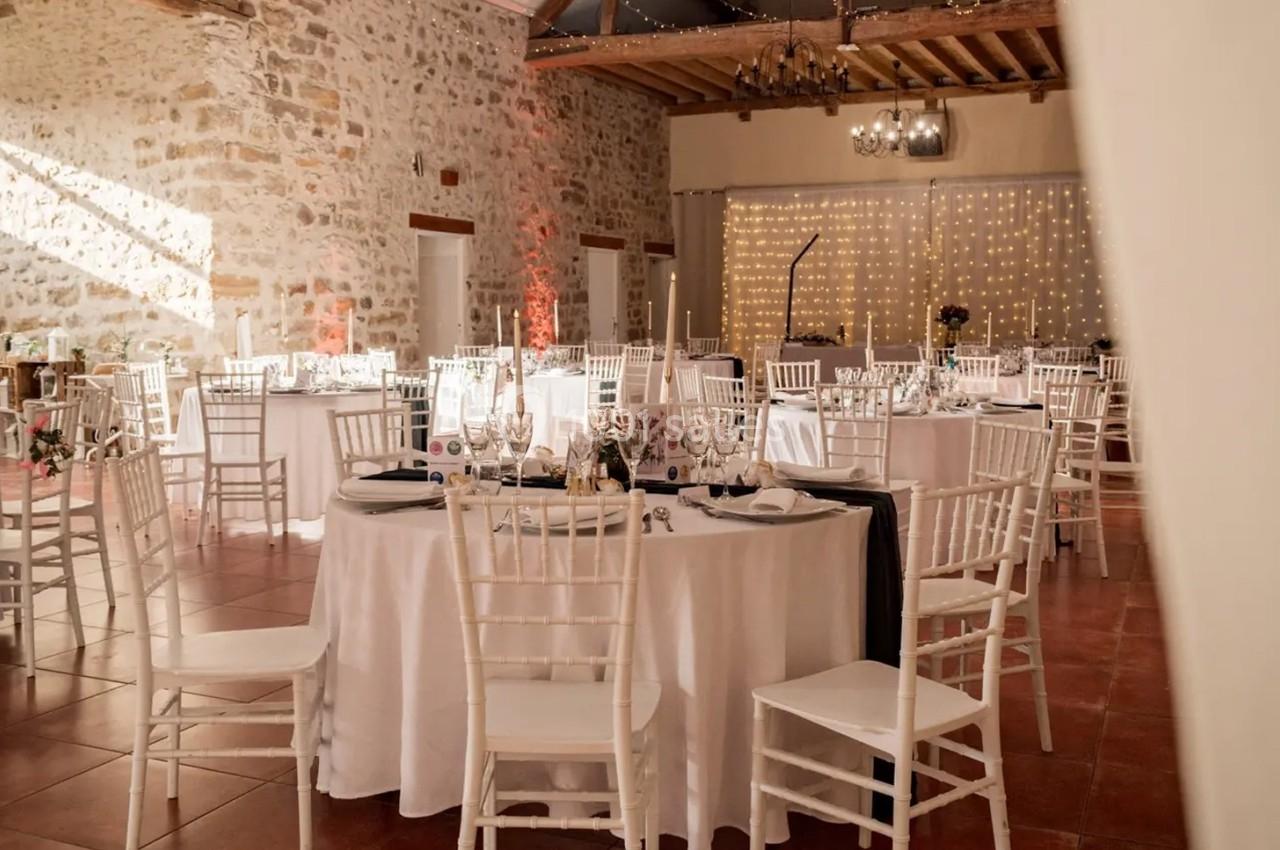 Location salle La Chapelle-Gauthier (Seine-et-Marne) - Nomade-Lodge #49 Salle de réception décorée avec des tables rondes dressées, chaises blanches et guirlandes lumineuses sur un mur en pierre.