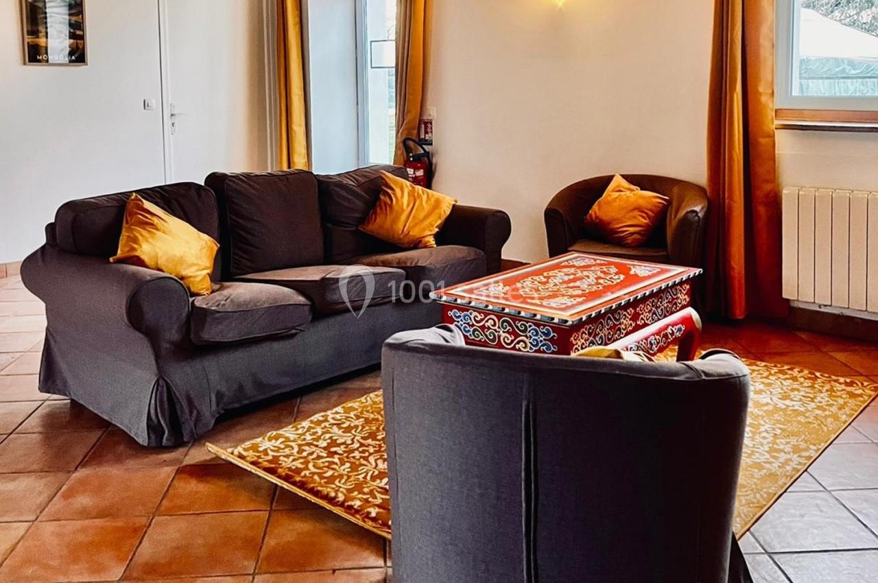 Location salle La Chapelle-Gauthier (Seine-et-Marne) - Nomade-Lodge #50 Salon avec canapé et fauteuils gris, coussins orange, table basse décorée et tapis assorti sur un sol en carrelage.