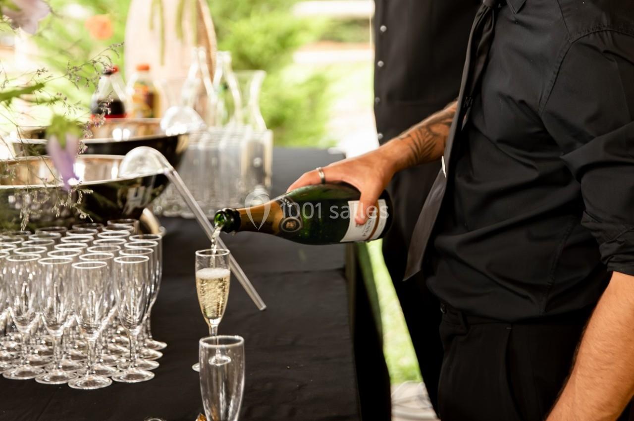 Location salle La Chapelle-Gauthier (Seine-et-Marne) - Nomade-Lodge #53 Un serveur verse du champagne dans une flûte sur une table garnie de verres et d'un seau à glace.
