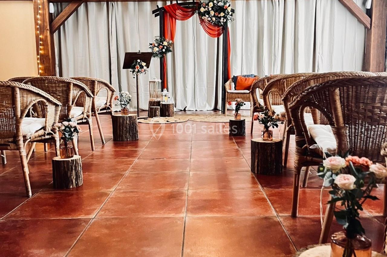 Location salle La Chapelle-Gauthier (Seine-et-Marne) - Nomade-Lodge #55 Disposition d'une salle de cérémonie avec des chaises en rotin, des fleurs et une arche décorée de tissu rouge et blanc.