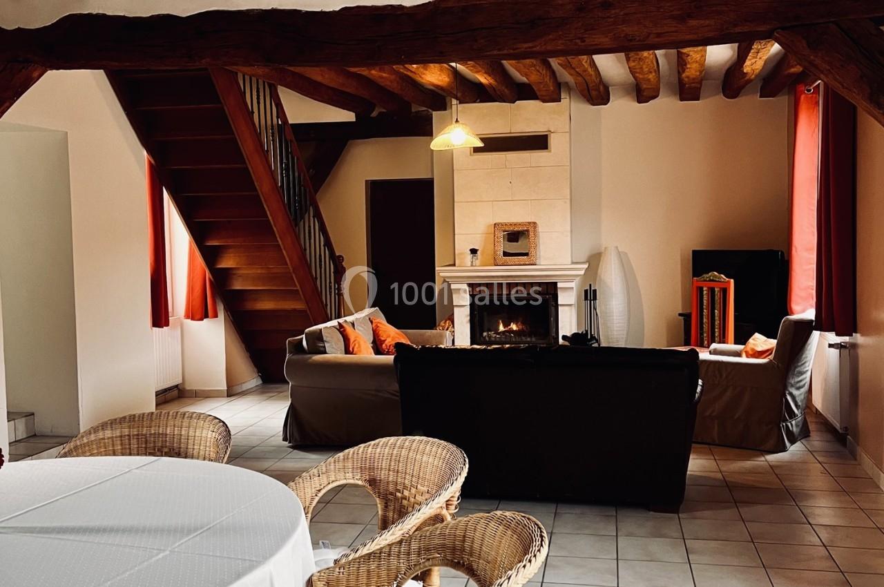 Location salle La Chapelle-Gauthier (Seine-et-Marne) - Nomade-Lodge #58 Salon chaleureux avec poutres apparentes, cheminée allumée, canapés, table en osier et escalier en bois.