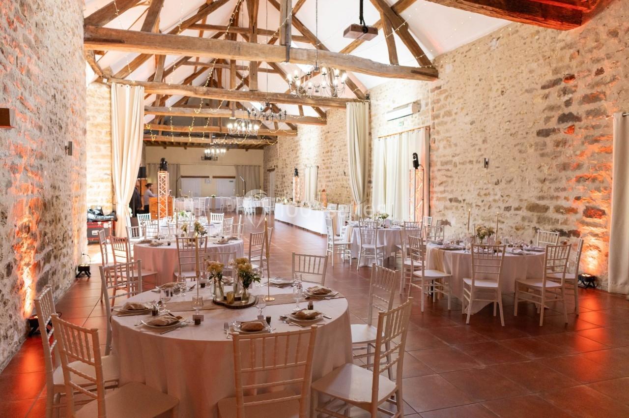 Location salle La Chapelle-Gauthier (Seine-et-Marne) - Nomade-Lodge #59 Salle de réception lumineuse avec murs en pierre, poutres apparentes et tables rondes décorées pour un événement.