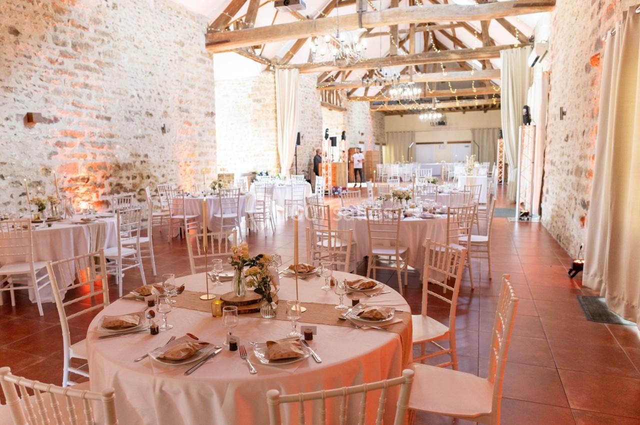 Location salle La Chapelle-Gauthier (Seine-et-Marne) - Nomade-Lodge #60 Salle de réception lumineuse avec murs en pierre, poutres apparentes et tables rondes dressées pour un événement.
