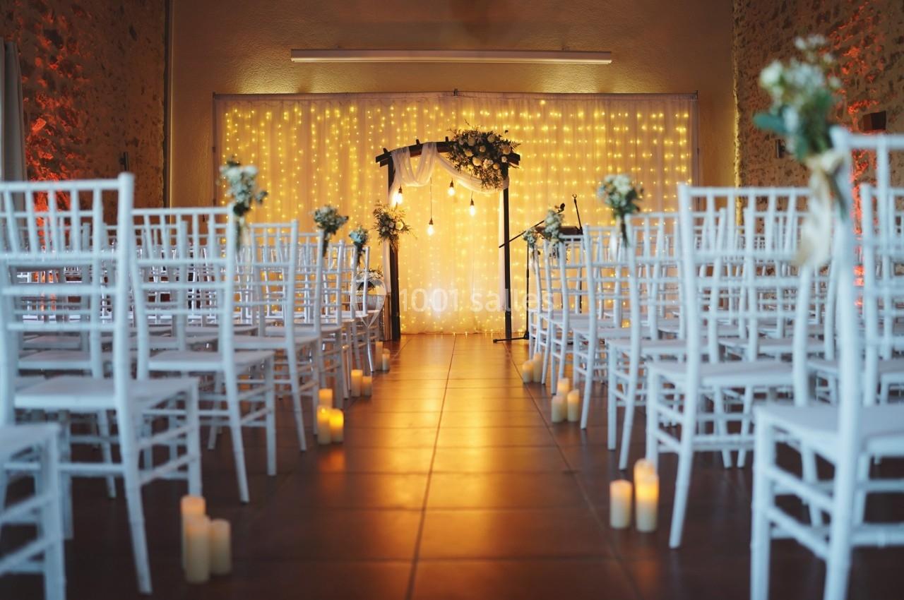 Location salle La Chapelle-Gauthier (Seine-et-Marne) - Nomade-Lodge #63 Allée centrale décorée de chaises blanches, bougies et guirlandes lumineuses menant à une arche florale.