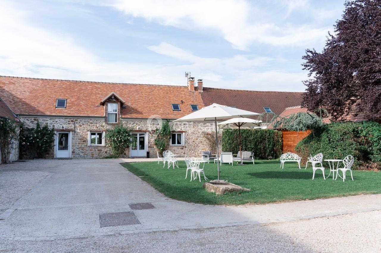 Location salle La Chapelle-Gauthier (Seine-et-Marne) - Nomade-Lodge #67 Cour d'une maison en pierre avec toit en tuiles, mobilier de jardin blanc et parasol sur une pelouse.