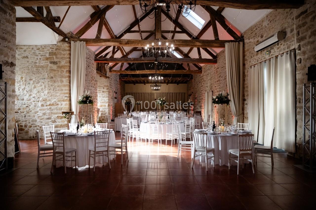 Location salle La Chapelle-Gauthier (Seine-et-Marne) - Nomade-Lodge #70 Salle de réception en pierres apparentes avec tables rondes dressées et décorations florales sous un plafond en poutres…