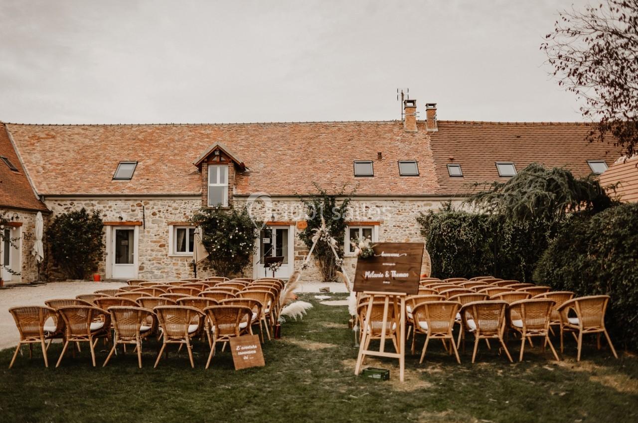 Location salle La Chapelle-Gauthier (Seine-et-Marne) - Nomade-Lodge #71 Chaises en rotin disposées en extérieur devant une maison en pierre pour une cérémonie avec arche décorée.