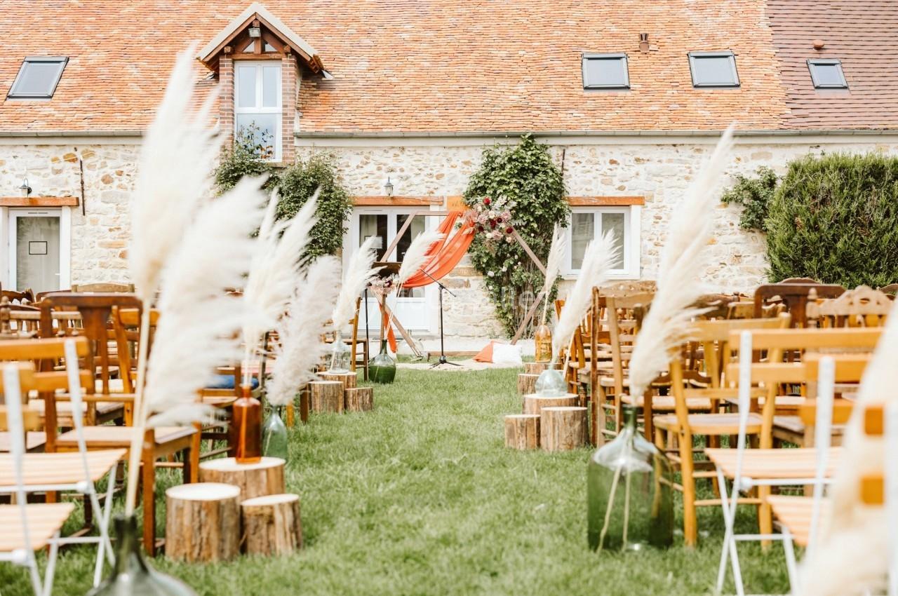 Location salle La Chapelle-Gauthier (Seine-et-Marne) - Nomade-Lodge #72 Allée centrale bordée de chaises et de décorations en pampas menant à une arche devant une maison en pierre.