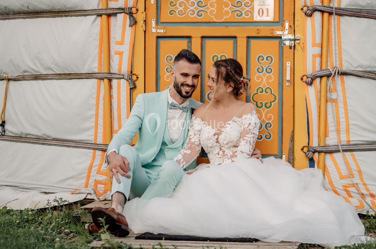 Un couple assis devant une yourte colorée, l'homme en costume turquoise et la femme en robe de mariée blanche.