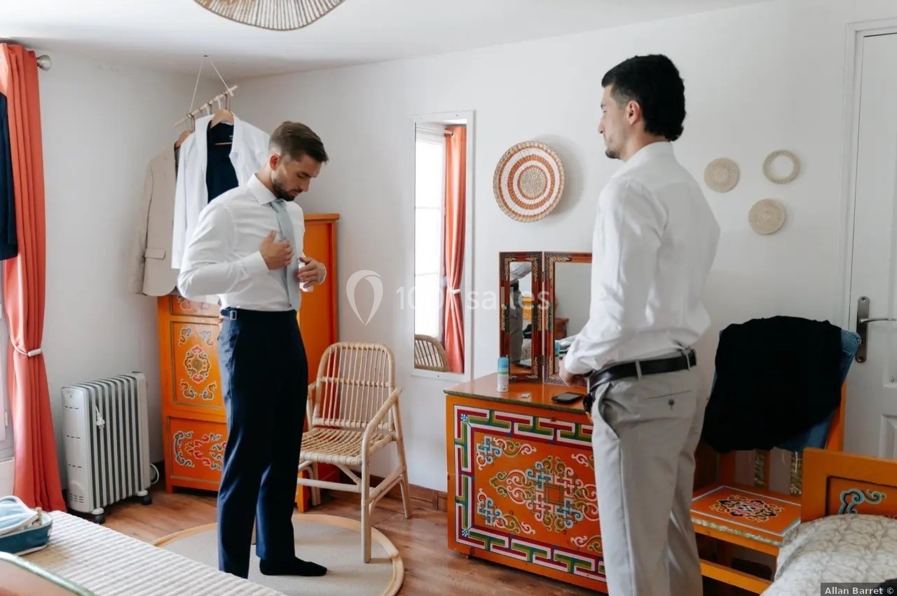 Deux hommes en chemise dans une chambre colorée, l'un ajustant son nœud papillon devant un miroir.