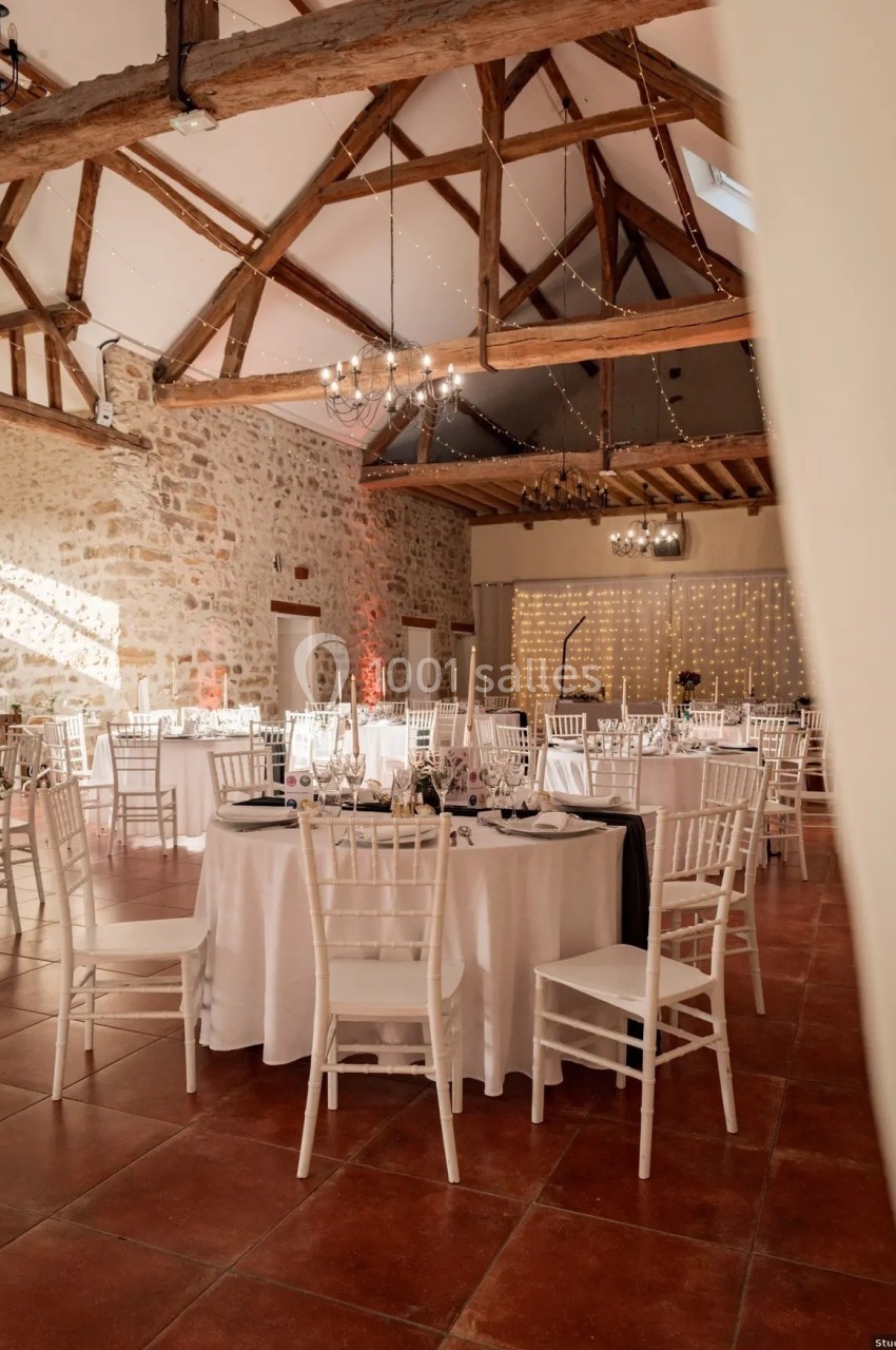 Salle de réception lumineuse avec tables rondes dressées, chaises blanches et poutres apparentes en bois.