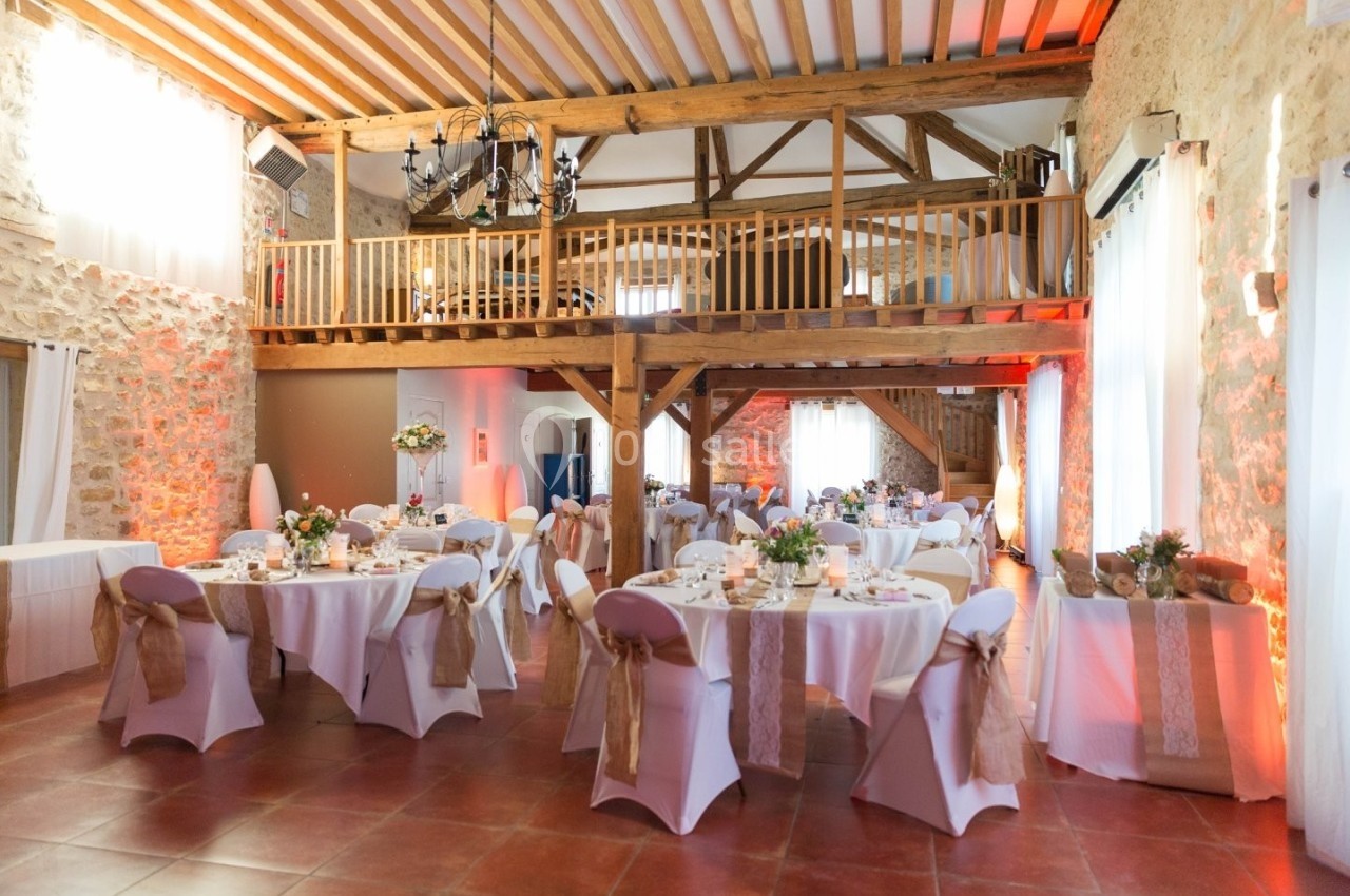 Salle de réception rustique avec poutres apparentes, tables décorées et chaises recouvertes de housses blanches.