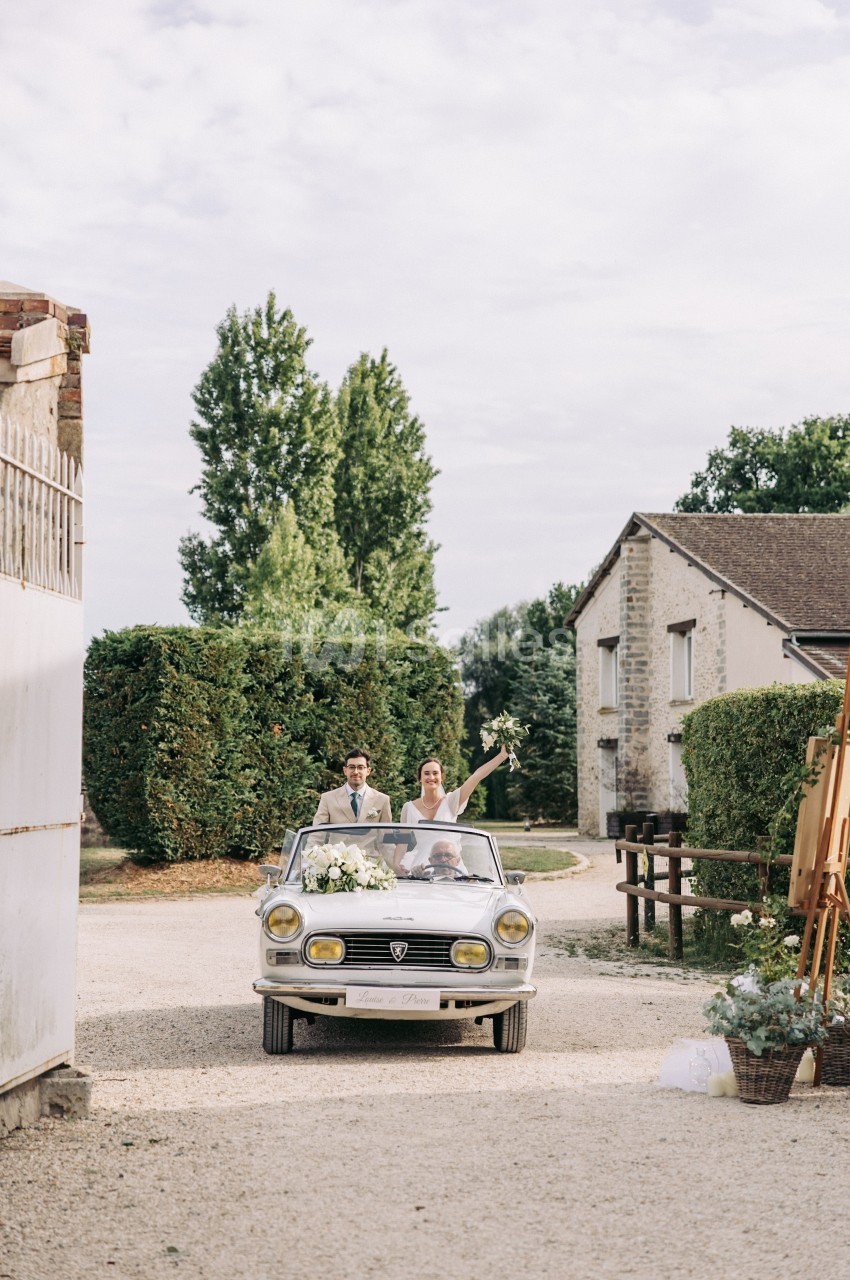 Un couple souriant dans une voiture décapotable ancienne, avançant sur une allée gravillonnée devant des bâtiments ruraux.