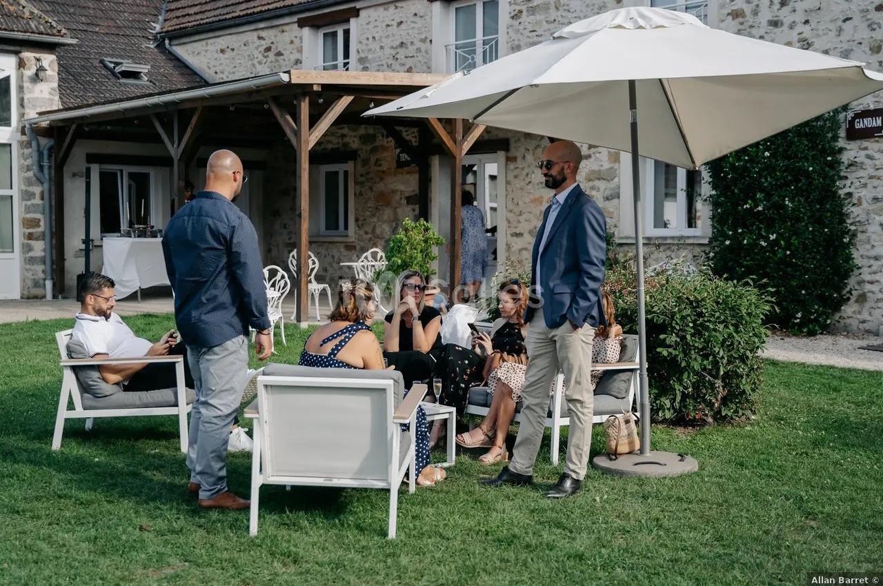 Des personnes discutent en plein air, assises et debout sous un parasol dans le jardin d'une maison en pierre.