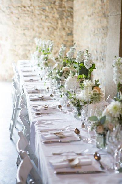Table longue décorée de nappes blanches, fleurs fraîches et vaisselle élégante dans une salle en pierre.