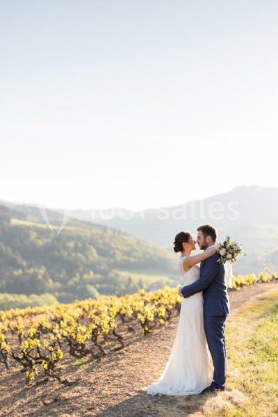 Un couple en tenue de mariage s'enlace dans un vignoble ensoleillé avec des collines verdoyantes en arrière-plan.