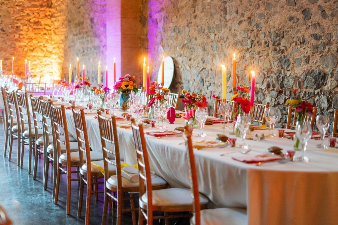 Grande table décorée pour un événement, avec nappes blanches, chandeliers allumés et arrangements floraux colorés.