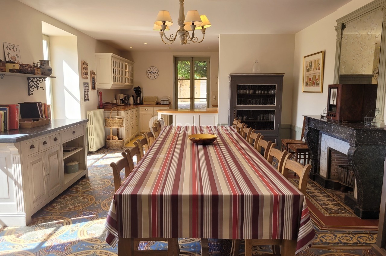 Grande cuisine lumineuse avec une longue table recouverte d'une nappe rayée, entourée de chaises en bois.