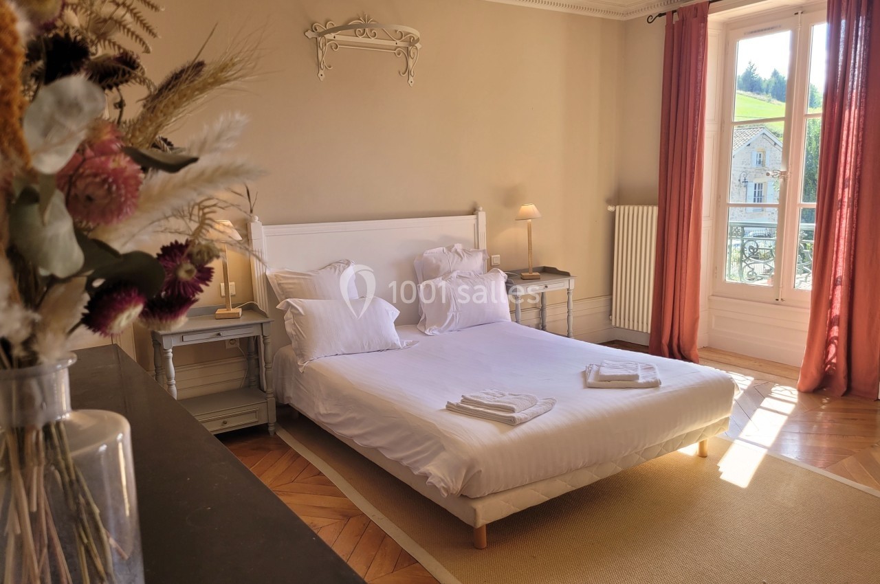 Chambre lumineuse avec lit double, draps blancs, table de chevet, bouquet de fleurs et fenêtre donnant sur l'extérieur.