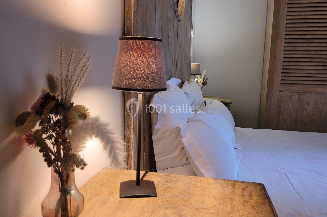 Lampe allumée sur une table en bois près d'un lit avec des coussins blancs et un vase de fleurs séchées.